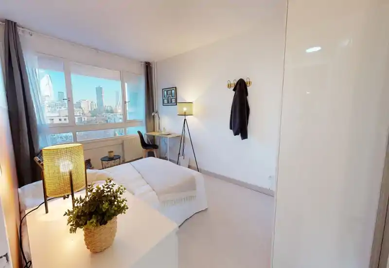 Chambre lumineuse et bien mise en scène avec lit double face à une grande fenêtre et un petit espace bureau ; linge neutre, lampes chaleureuses et plante apportent une ambiance cosy.