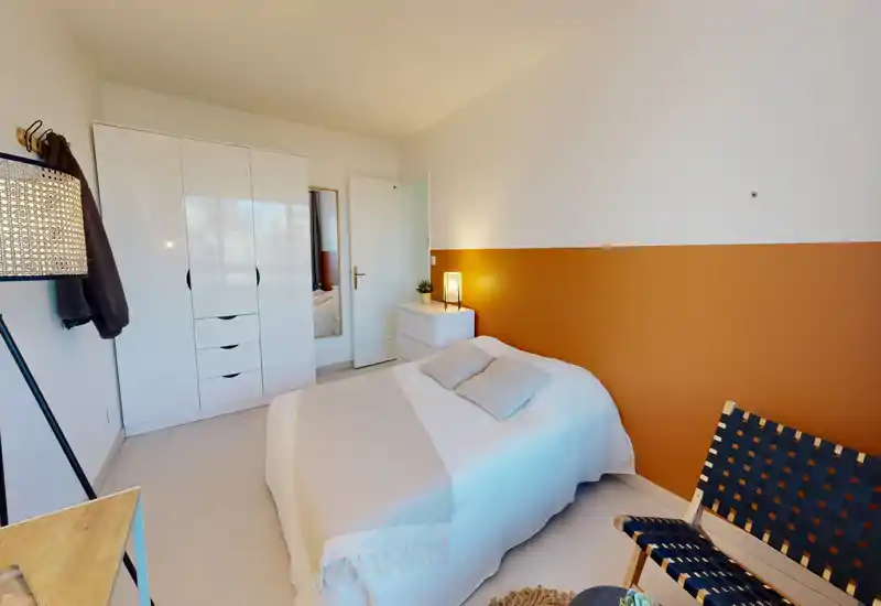 Chambre moderne et lumineuse avec un lit double confortable, mur d'accent chaleureux et armoire blanche. Textiles doux, lampe de chevet et décoration soignée donnent une atmosphère accueillante, idéale pour une annonce.
