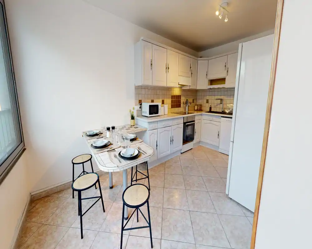 Cuisine lumineuse et compacte avec meubles blancs, plan de travail d'angle, électroménagers modernes et petite table avec tabourets — idéale pour un appartement chaleureux.