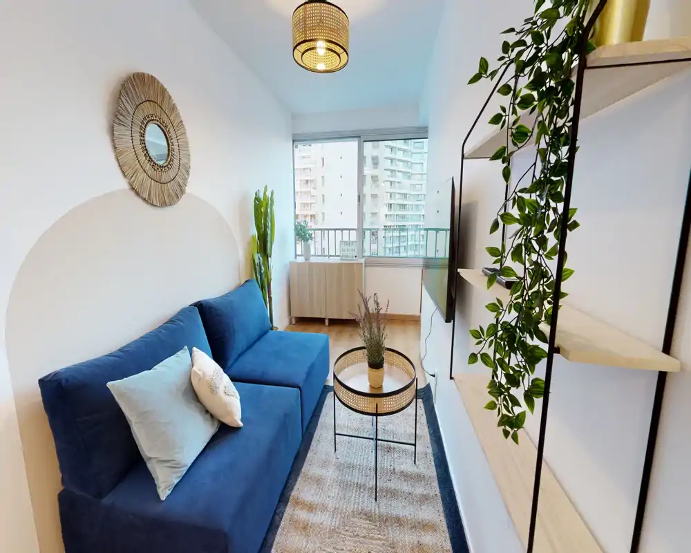 Salon lumineux et élégant avec un canapé bleu, tapis texturé et une petite table basse. Grande fenêtre offrant une vue sur la ville et lumière naturelle ; étagères décoratives et plantes ajoutent une touche cosy et moderne.