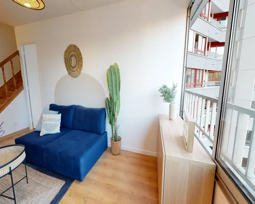 Coin salon lumineux et soigné avec un canapé bleu foncé, coussins décoratifs, plantes et une grande fenêtre apportant une belle lumière naturelle — idéal pour les annonces.