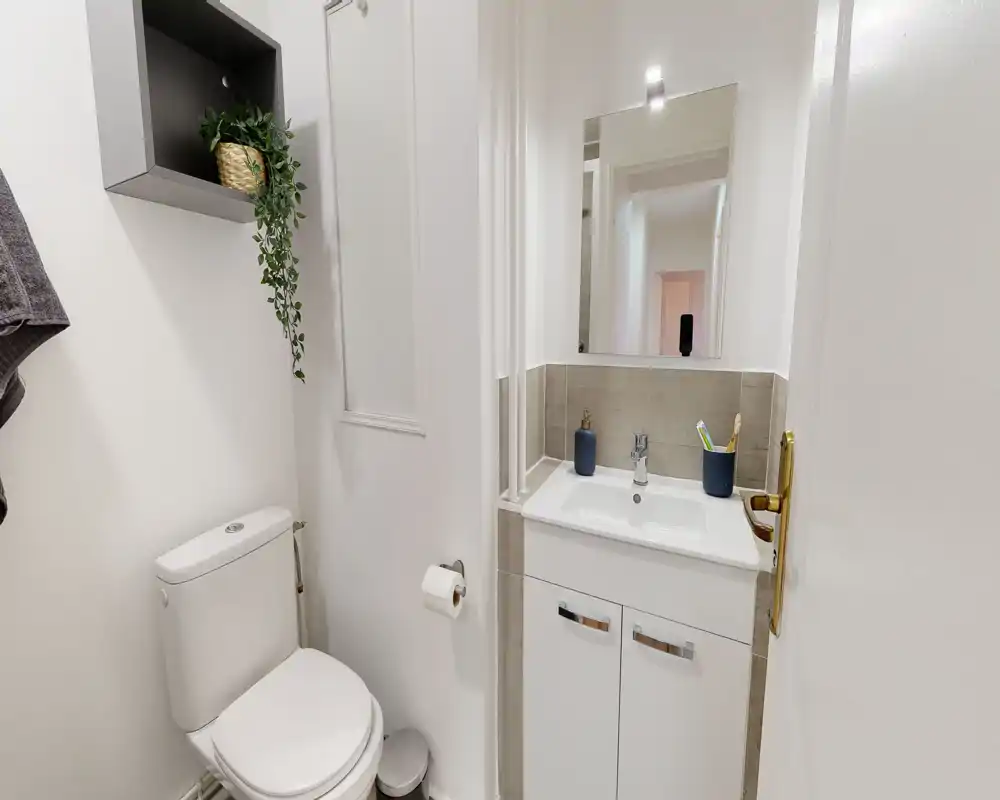 Salle de bain compacte et moderne avec toilette, meuble-vasque avec rangement, miroir et accessoires soignés. Murs blancs et carrelage clair pour une apparence propre et minimaliste adaptée aux annonces.