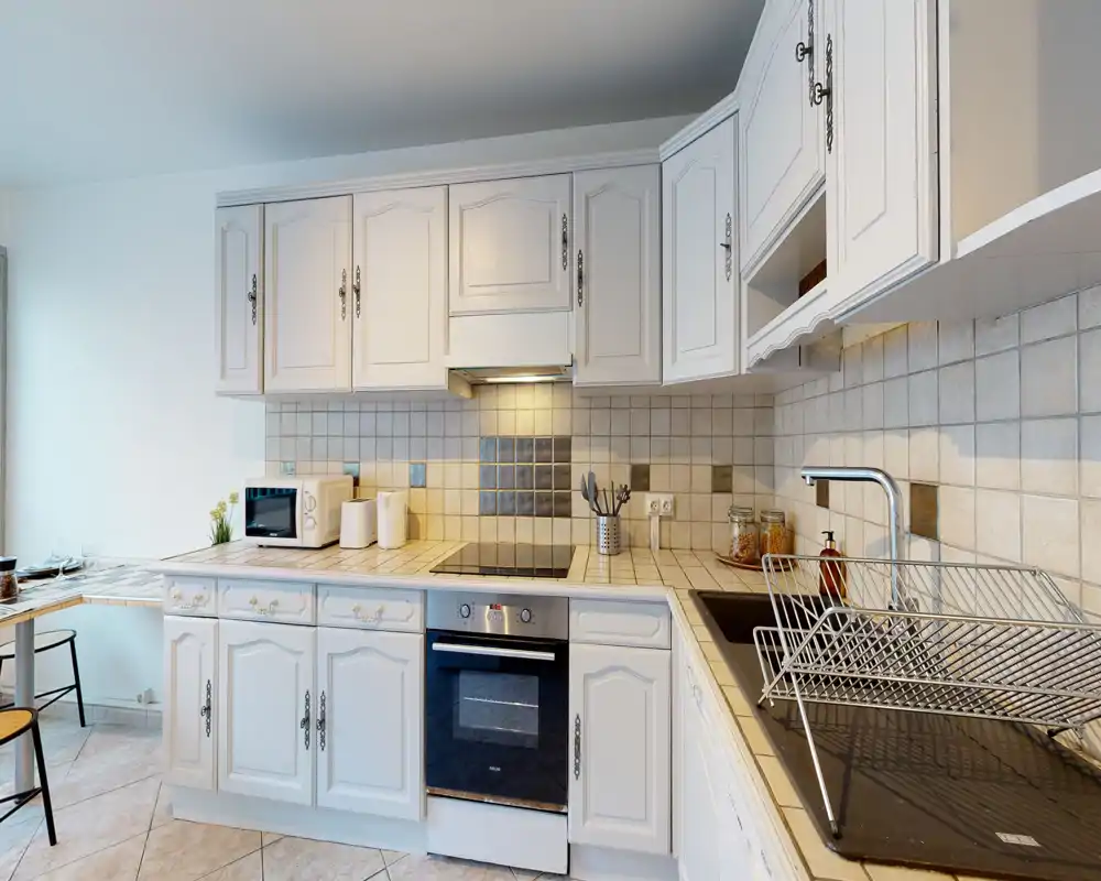 Cuisine lumineuse et bien équipée avec meubles blancs, crédence carrelée, four encastré et plaque à induction — un espace propre et convivial idéal pour cuisiner.