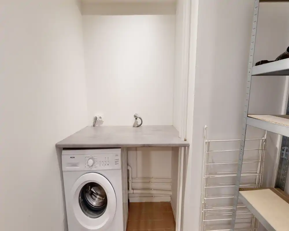 Petit espace buanderie/utility avec lave-linge frontal sous un plan de travail et robinets muraux au-dessus ; étagères de rangement visibles à droite.