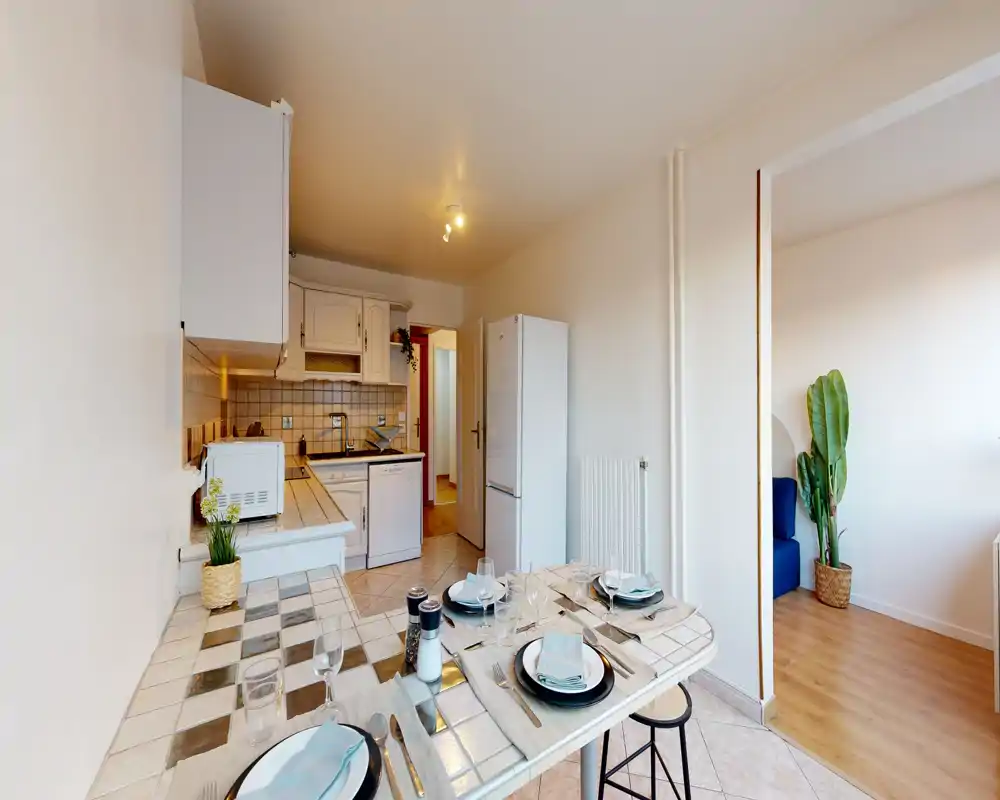 Kitchenette lumineuse avec un comptoir carrelé dressé pour le repas, meubles blancs, réfrigérateur et micro-ondes. L’espace compact est mis en scène de façon chaleureuse et s’ouvre sur le salon.