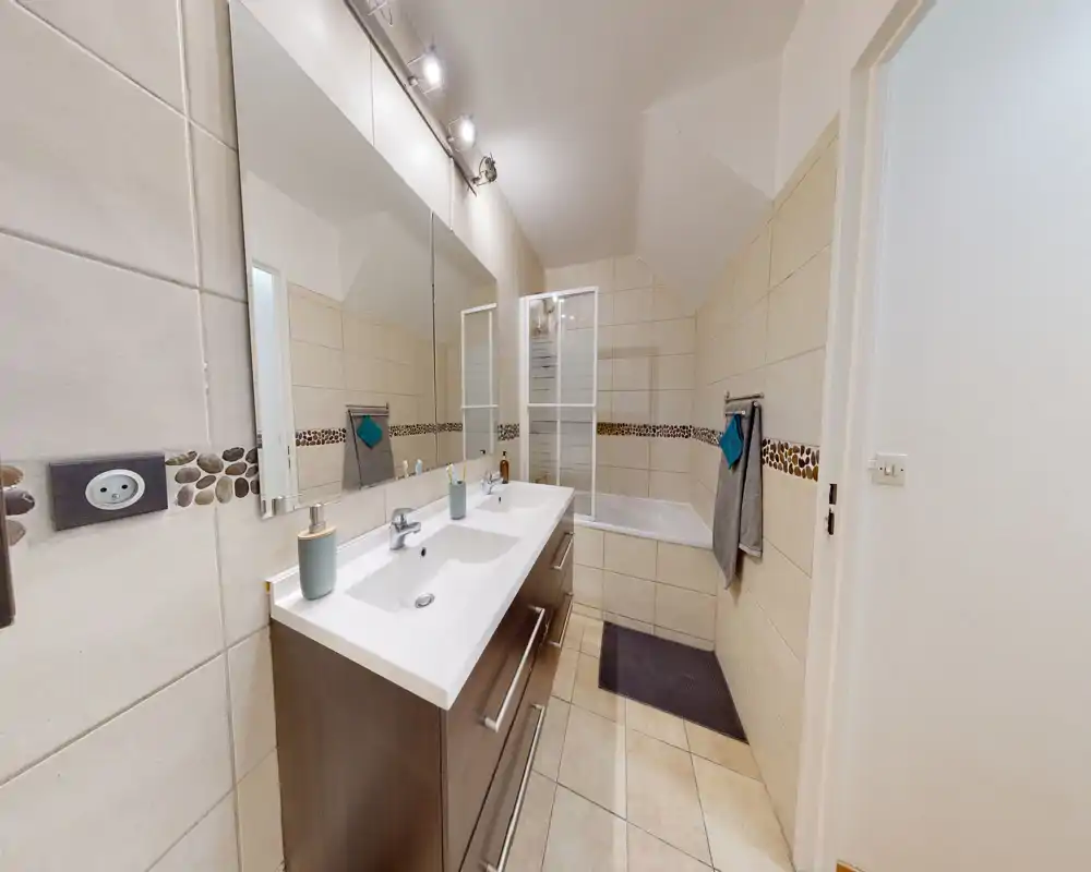 Salle de bain moderne et bien éclairée avec meuble vasque double, grand miroir, baignoire avec paroi de douche pliante en verre et carrelage au sol et aux murs.