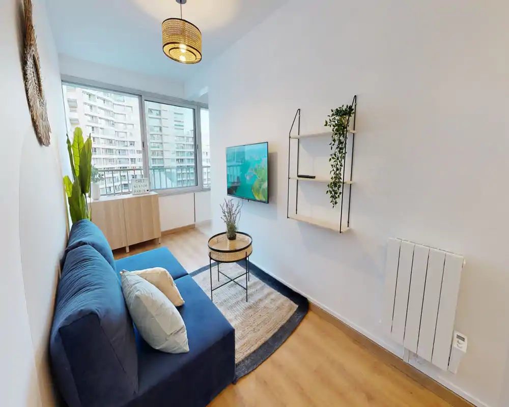 Petit salon lumineux avec canapé bleu, coussins décoratifs, petite table basse et TV murale ; de grandes fenêtres apportent la lumière naturelle et un tapis chaleureux unifie l'espace.