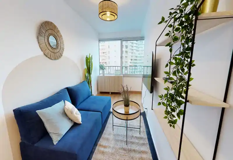 Salon lumineux et élégant avec un canapé bleu, tapis texturé et une petite table basse. Grande fenêtre offrant une vue sur la ville et lumière naturelle ; étagères décoratives et plantes ajoutent une touche cosy et moderne.