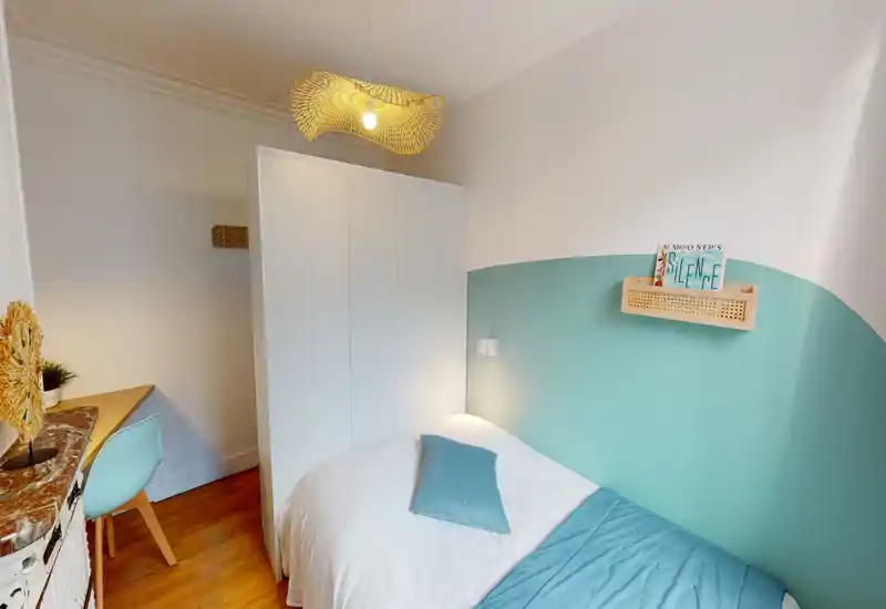 Camera da letto compatta e ben arredata con letto matrimoniale, parete accentata turchese e luce naturale morbida. Piccolo scrittoio e armadio la rendono pratica per una persona o soggiorni brevi.