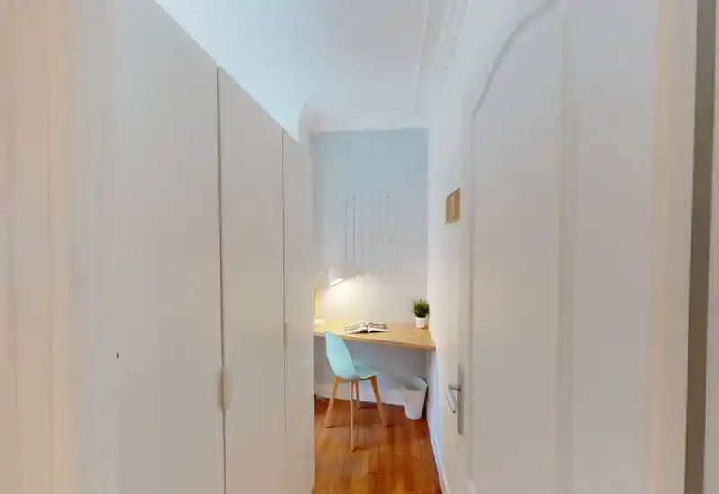 Piccolo angolo luminoso con scrivania compatta, sedia moderna e armadio a muro; pavimento in legno e lampada da scrivania creano un ambiente accogliente e funzionale nell'angolo della stanza.