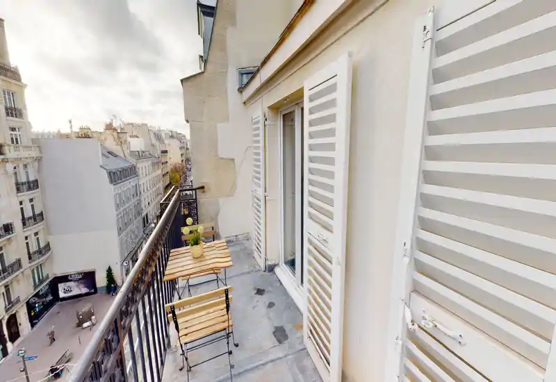 Balcone parigino soleggiato e stretto con un piccolo tavolo bistro in legno e due sedie pieghevoli, affaccio su una strada cittadina e facciate in stile Haussmann — perfetto per il caffè mattutino.