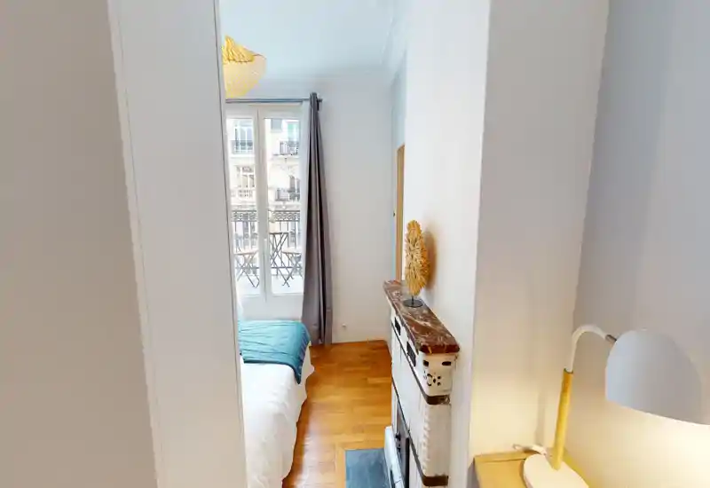 Camera da letto luminosa e compatta con letto ordinato, pavimento in parquet e finestra su un piccolo balcone. Tessili morbidi e illuminazione calda rendono l'ambiente accogliente.