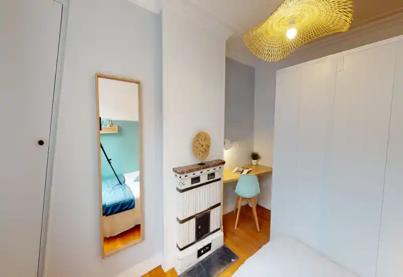 Angolo camera compatto e ben illuminato con vista parziale del letto, armadio a muro, una scrivania piccola con sedia e un antico riscaldatore decorativo. L’ambiente è accogliente con pavimento in legno e luce calda.