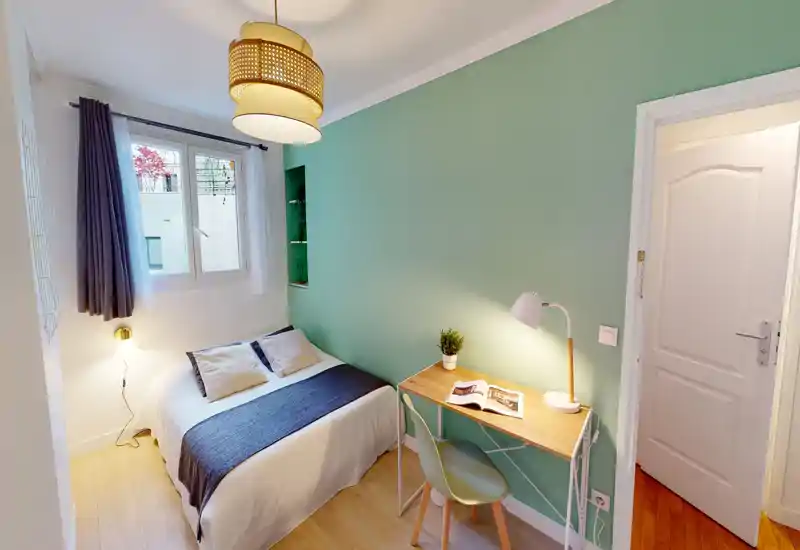 Chambre compacte et bien aménagée avec lit double, lampes de chevet et petit bureau devant un mur vert menthe ; la lumière de la fenêtre et les lampes chaudes créent une atmosphère cosy.