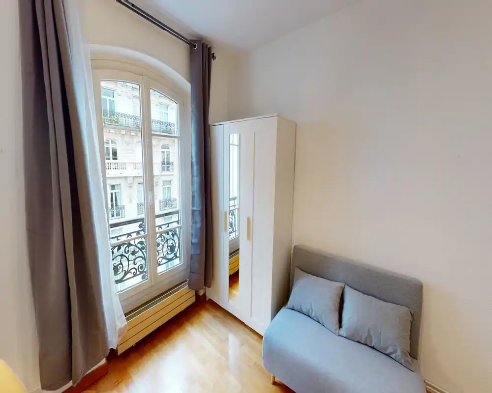 Petite chambre avec grande fenêtre cintrée et balcon, parquet clair, armoire blanche avec miroir et un petit canapé rembourré pouvant servir de couchage d'appoint.