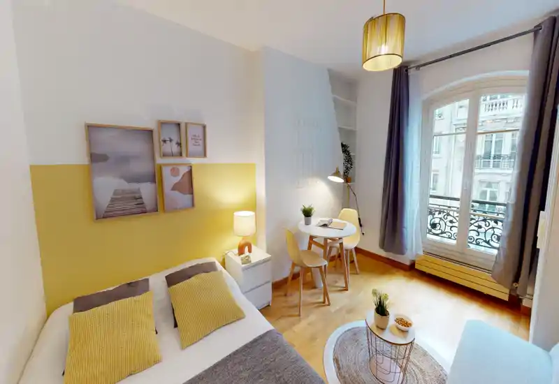 Bright, styled bedroom with a double bed, bedside table and lamp, small dining/work table by a tall arched window letting in natural light. Soft textiles, framed wall art and warm yellow accents create a cozy, modern feel ideal for rental photos.