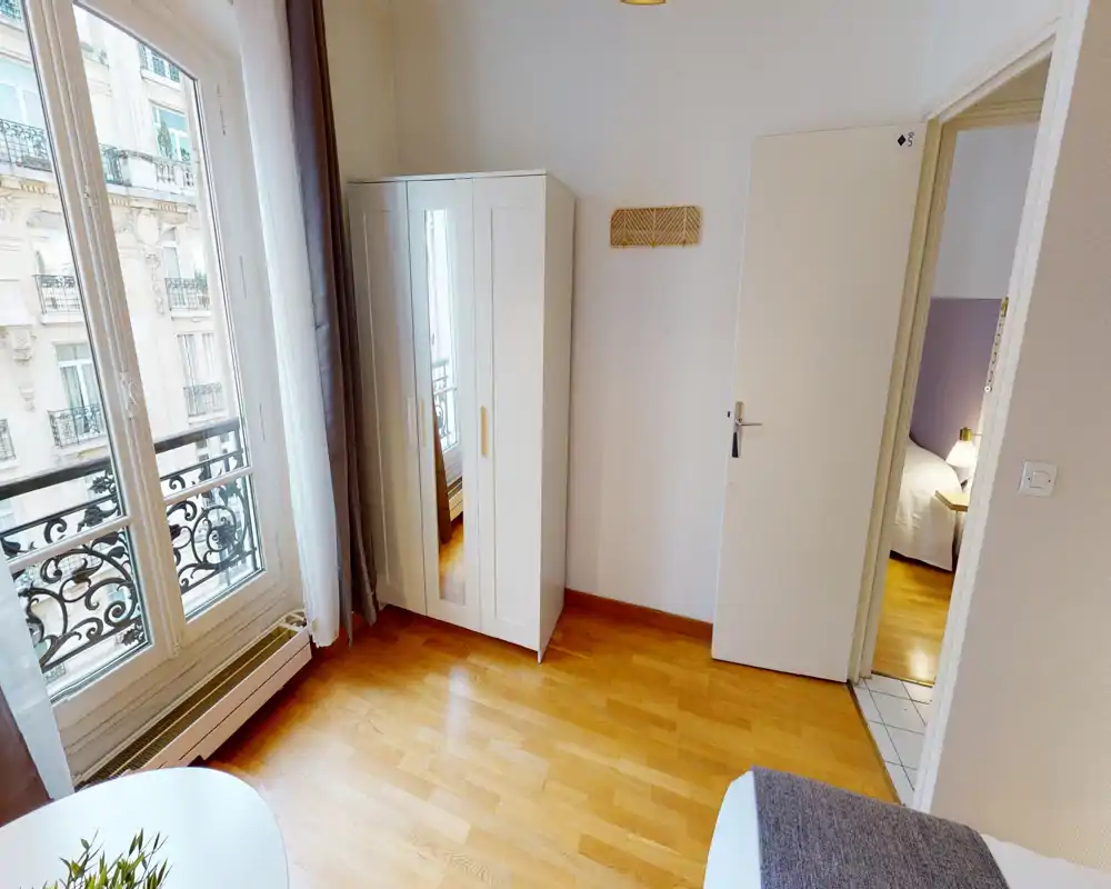 Angolo di camera luminoso e compatto con armadio con specchio, ampia finestra in stile parigino e vista parziale del letto. La luce naturale illumina la stanza e il pavimento in legno dona calore e pulizia.