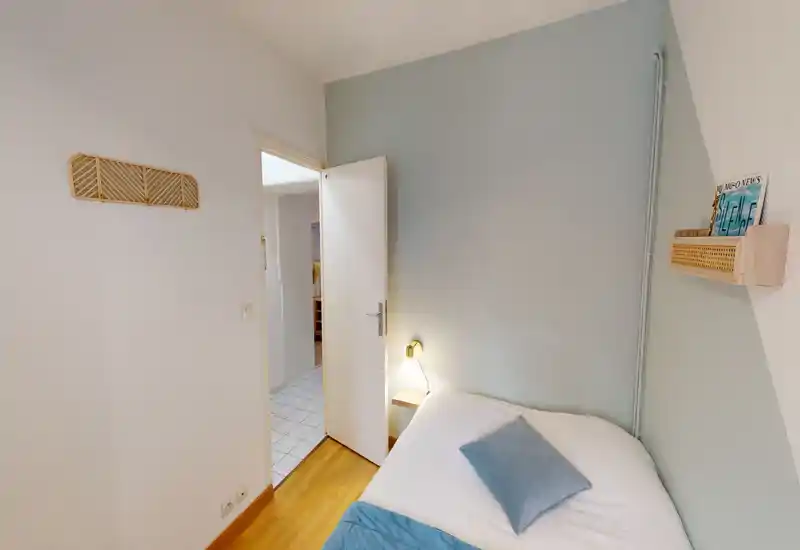 Small cozy bedroom with a single bed dressed in white linens and blue accents, a bedside lamp and soft, neutral walls that create a calm atmosphere.