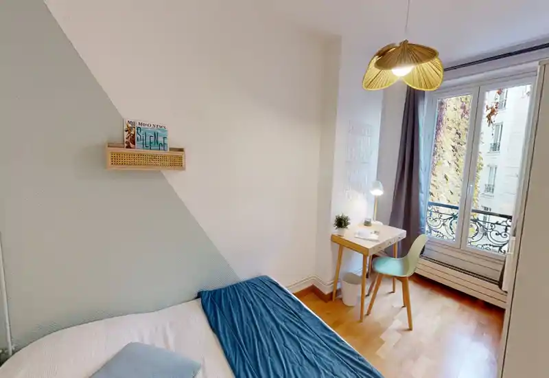 Bright, compact bedroom with a comfortable bed, desk by the window and natural light from a Parisian-style balcony. Soft textiles and minimal decor create a calm, inviting atmosphere ideal for short stays.