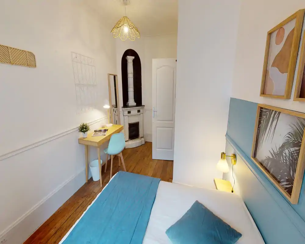 Chambre lumineuse et stylée avec lit double habillé de textiles bleu-vert, lampe de chevet, cadres muraux et un petit bureau avec chaise — espace chaleureux et bien éclairé, parfait pour une ou deux personnes.