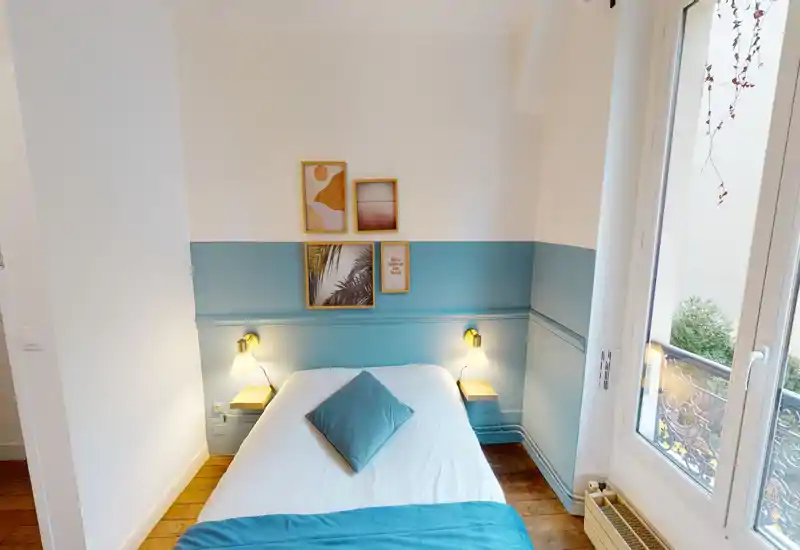 Chambre lumineuse et chaleureuse avec un lit double bien fait, mur d'accent bleu-vert, lampes de chevet et tableaux encadrés. Lumière naturelle provenant d'une grande fenêtre donnant sur un petit balcon.