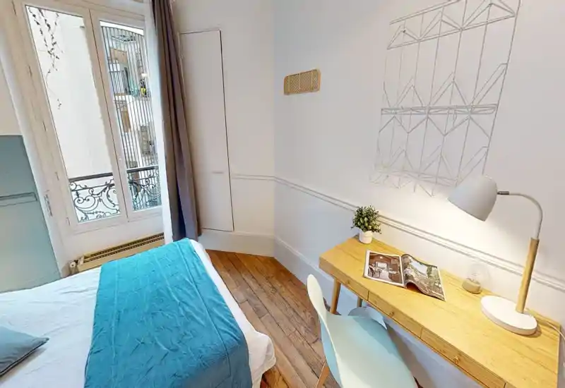 Chambre lumineuse et élégante avec un lit confortable, parquet et un petit bureau près de la fenêtre. La lumière naturelle et la décoration apaisante offrent une atmosphère cosy.