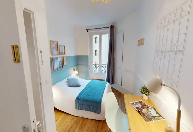 Chambre lumineuse et soignée avec lit double en linge blanc et touches turquoise, sol en bois et un petit bureau contre le mur — idéale pour un séjour confortable.