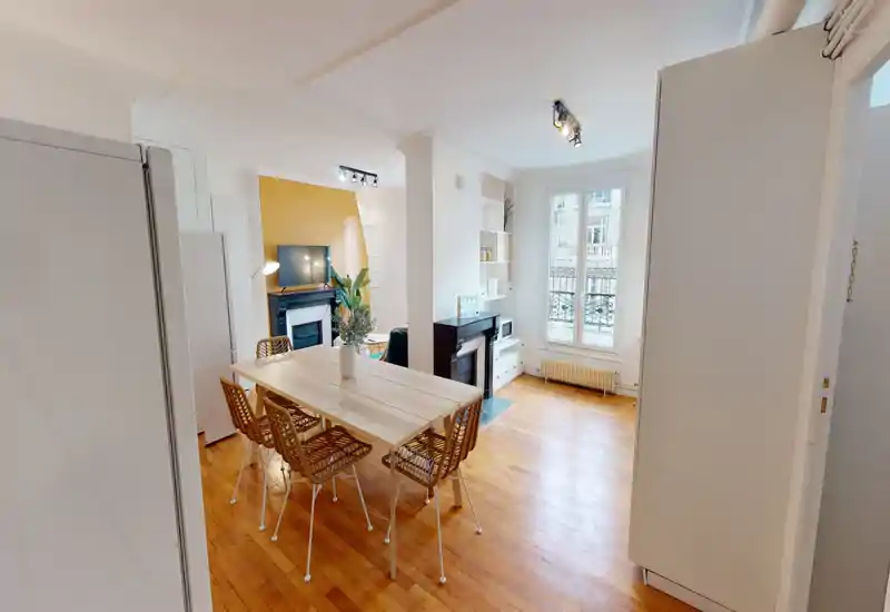Bright, modern dining area with a wooden table and woven chairs, open to a living corner with a fireplace and large window providing natural light.