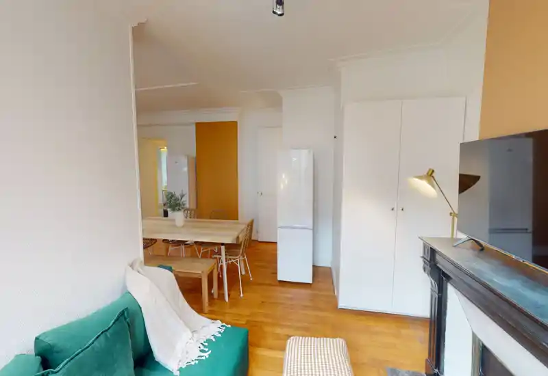 Spacious, well-lit living area with a green sofa, wooden floors and a dining table visible in the background. The room blends sitting and dining spaces with warm tones and modern, cozy styling.