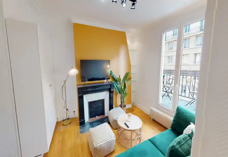 Séjour lumineux et élégant avec canapé vert, poufs, table basse et télévision au-dessus d'une cheminée décorative ; grandes fenêtres donnant sur un petit balcon.