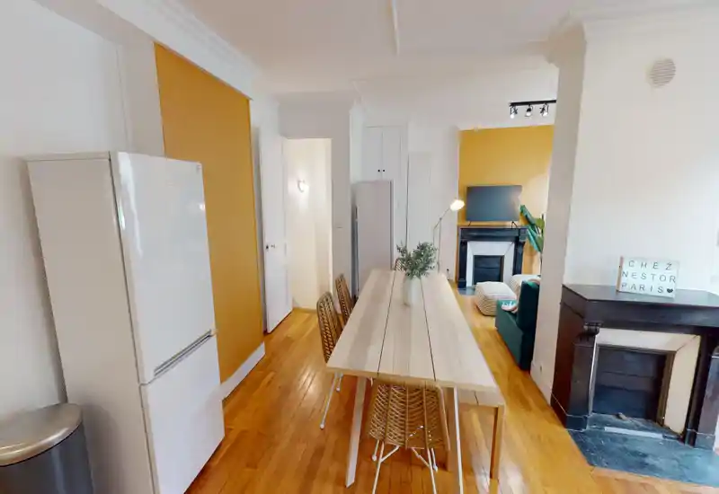 Open-plan dining area with a central wooden table and woven chairs, warm yellow accent walls and visible living area beyond. Bright, cozy atmosphere ideal for meals and socializing.