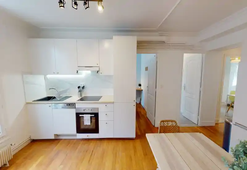 Modern compact kitchen with white cabinets, integrated oven and dishwasher, warm wooden floors and a small dining table—bright, clean and ideal for a city apartment.