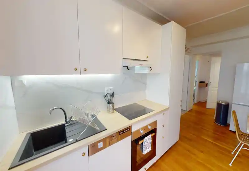 Modern, compact kitchen with a built-in oven and induction hob, stainless sink, under-cabinet lighting and white cabinetry, set on warm wooden floors.