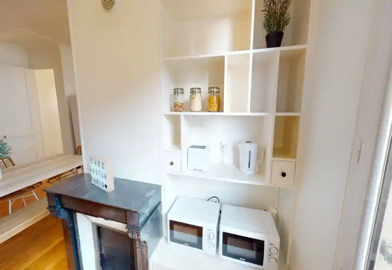 Compact kitchenette with white shelving, two countertop microwaves, a kettle and toaster, and decorative jars — bright and neatly arranged.