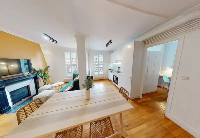 Bright, styled open-plan living area with a dining table in the foreground, a comfortable sofa and a TV by a decorative fireplace; an integrated kitchen runs along the right wall.