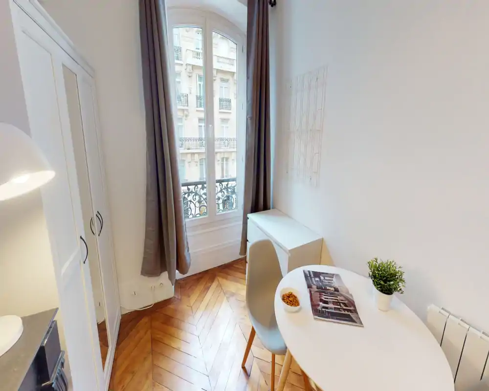 Espace de vie lumineux et compact avec table ronde, chaise, bureau près de la fenêtre et armoire. Parquet en chevrons et grande fenêtre avec rideaux donnent une ambiance chaleureuse, typique parisienne.