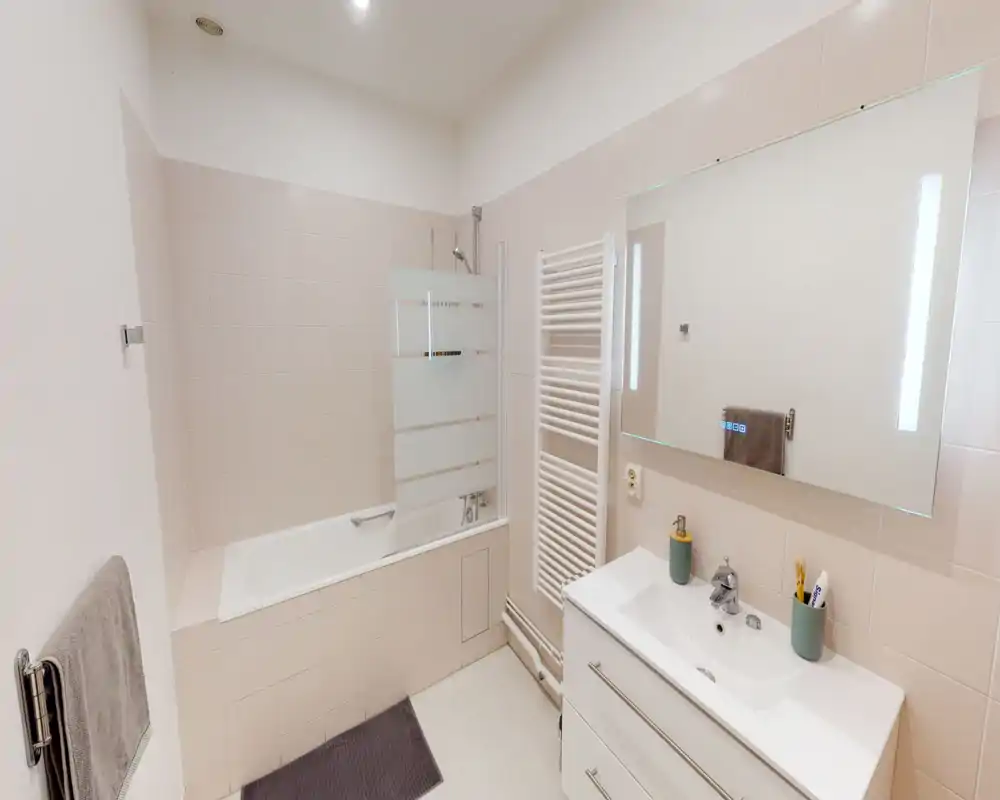 Salle de bains moderne et lumineuse avec baignoire-douche, grand miroir, radiateur porte-serviettes et meuble lavabo blanc avec rangements.