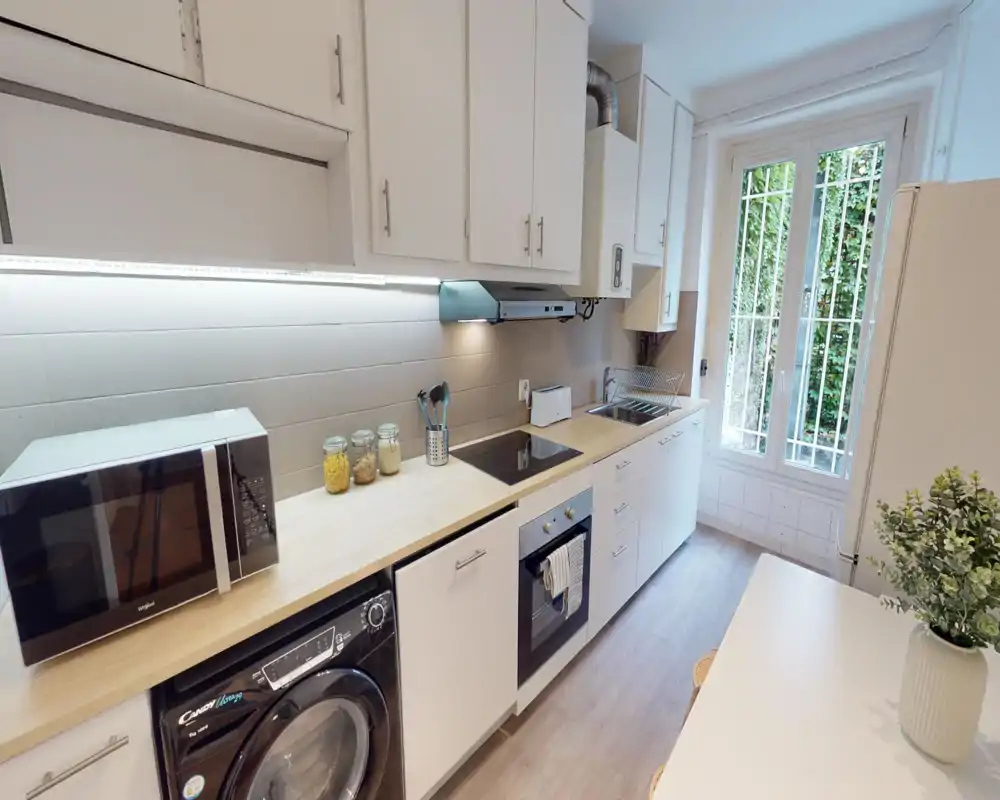 Cuisine moderne en longueur lumineuse avec plans de travail en bois clair, placards blancs, four encastré et plaque à induction, micro-ondes, évier près d'une grande fenêtre et bocaux décoratifs.