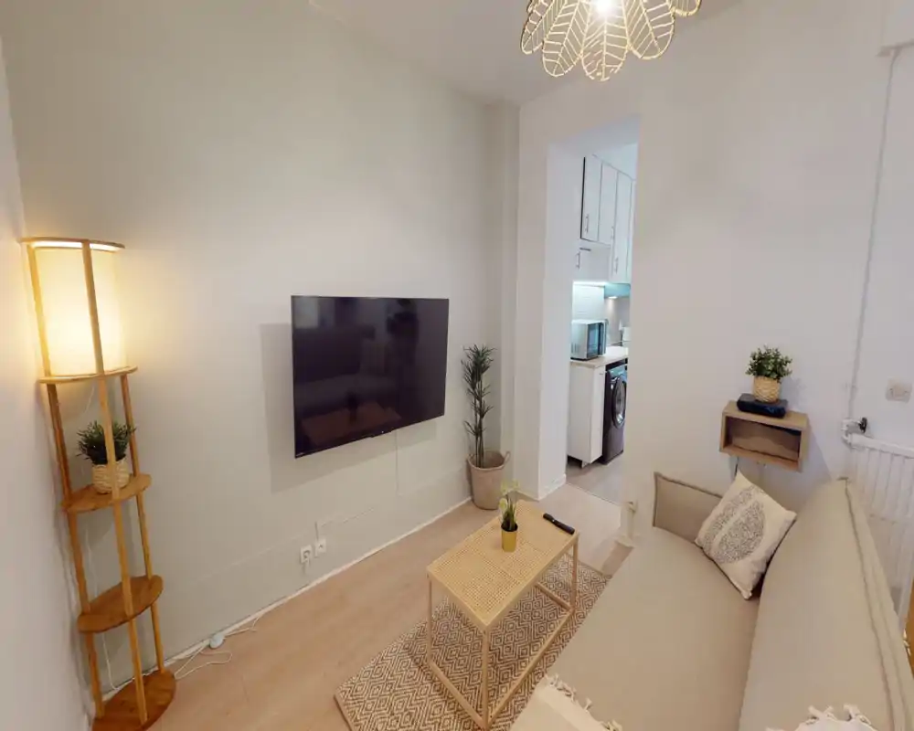 Salon chaleureux et bien mis en scène avec TV murale, canapé confortable, table basse en rotin et éclairage doux — idéal pour se détendre.