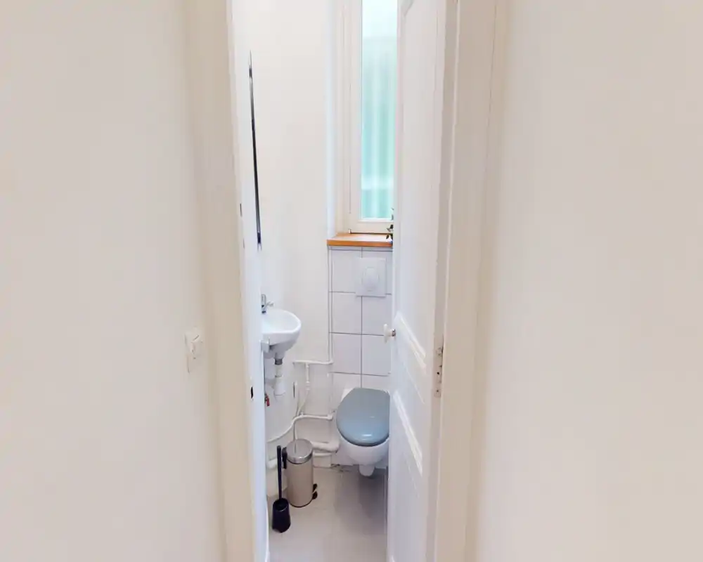 Salle de bain compacte et lumineuse avec WC suspendu et petit lavabo d'angle. Carrelage neutre et fenêtre dépoli pour l'intimité et la lumière naturelle.