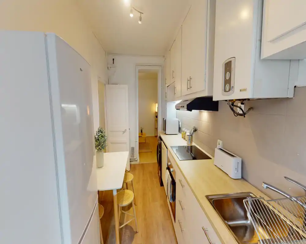 Cuisine en couloir lumineuse avec plans de travail en bois, placards blancs et appareils intégrés. Une petite table avec des tabourets et une plante décorative apportent une atmosphère cosy et fonctionnelle.