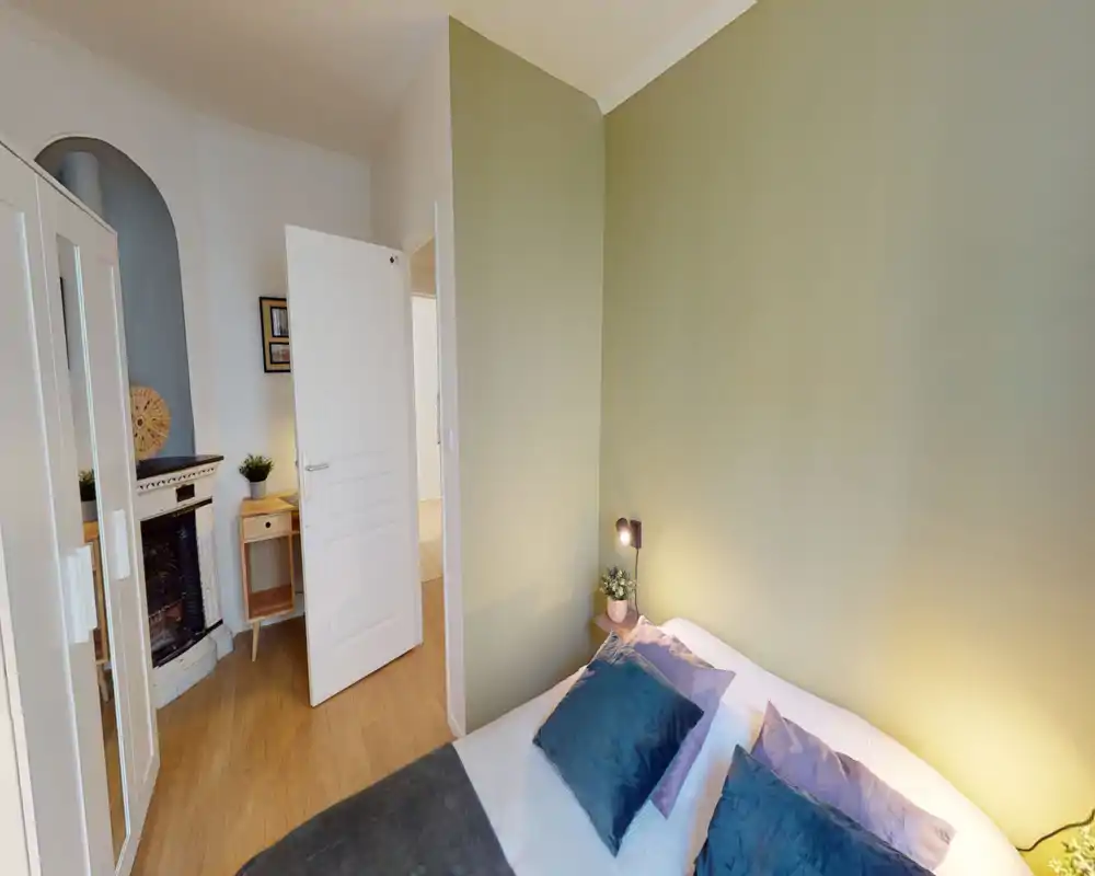 Chambre lumineuse et chaleureuse avec un lit double bien fait, coussins décoratifs et lampe de chevet. Mur vert neutre et sol en bois pour une ambiance conviviale.