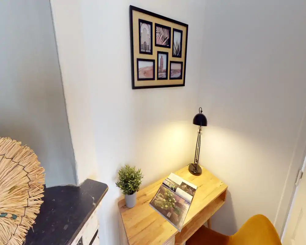 Un coin salon chaleureux avec un bureau en bois, une lampe de table à lumière chaude, des cadres au mur et une petite plante — parfait pour lire ou travailler confortablement.