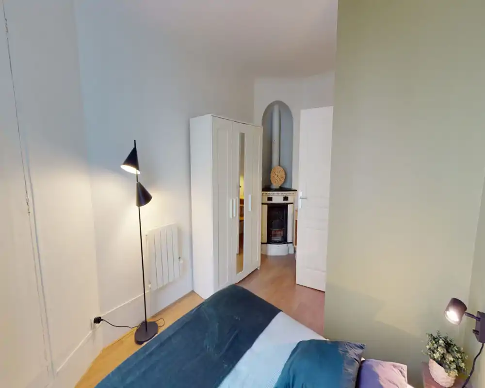 Coin chambre lumineux et soigné avec un lit double partiellement visible, une lampe sur pied, une armoire à portes miroir et une cheminée décorative dans une alcôve en arc.