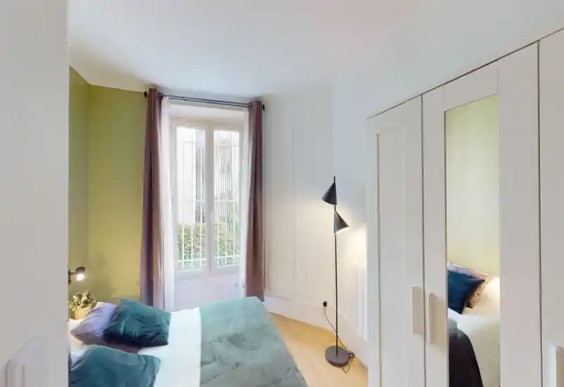 Bright, stylish bedroom with a double bed dressed in green textiles, a floor lamp and a window with curtains letting in natural light. The room features a wardrobe with mirrored door and warm wooden floors, creating a welcoming atmosphere for guests.
