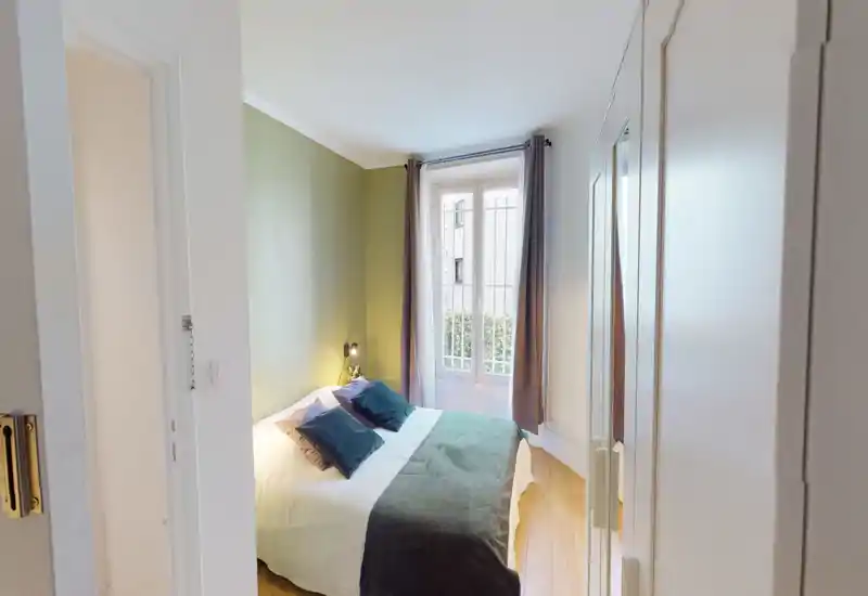 Bright, cozy double bedroom with a neatly made bed, soft pillows and a large window letting in natural light. Neutral wood flooring and green accent wall create a calm, stylish atmosphere ideal for relaxing stays.