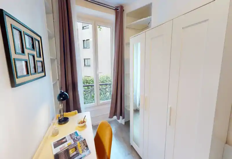 Small bedroom corner with a workspace: a light wood desk and mustard chair face a tall window with curtains, and a large white wardrobe provides storage. The room feels tidy, well lit and suitable for a single occupant.