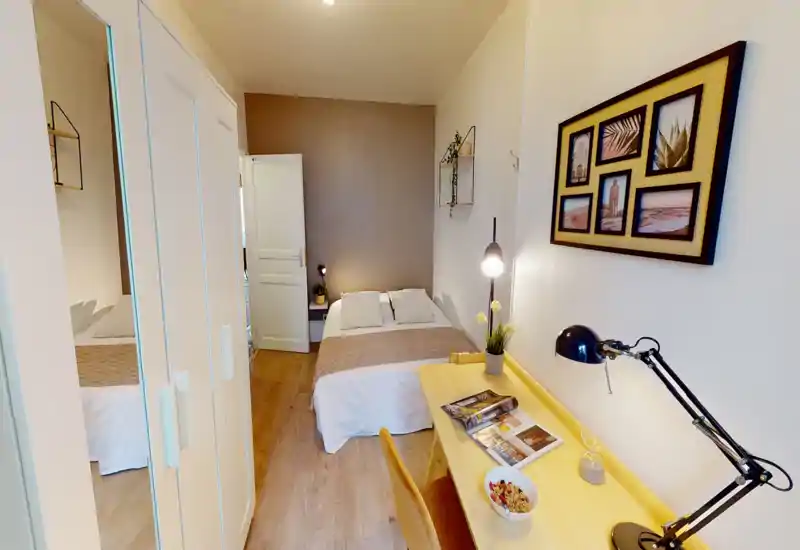 Bright, cozy bedroom with a neatly made double bed centered against a warm accent wall, a desk with task lamp in the foreground and a wardrobe with mirrored doors to the left.