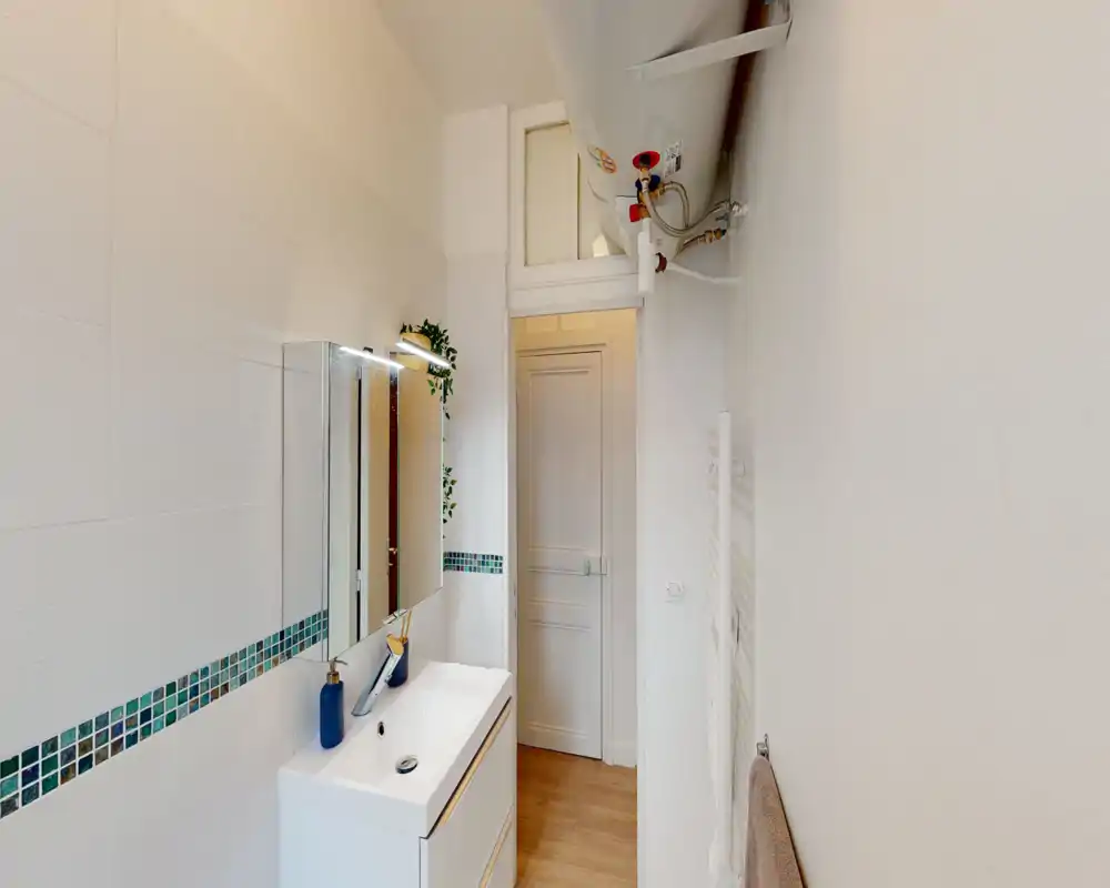 Compact, modern bathroom with a white vanity, sink, mirror cabinet and decorative tile border. Bright, clean finish and wooden-look floor visible through the doorway.