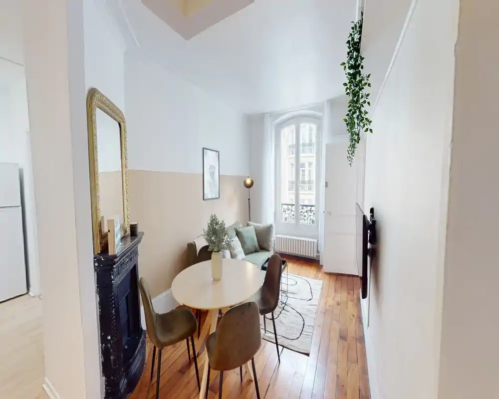 Bright, well-staged living room with a compact dining table, green sofa, decorative rug and large window providing natural light — ideal for a cozy city apartment listing.